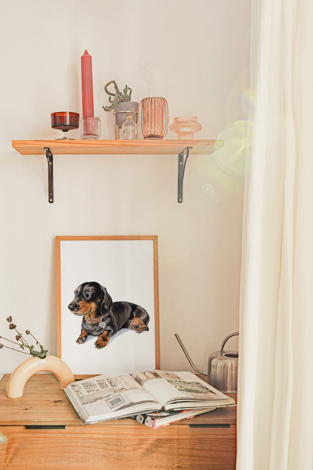 A framed custom pet portrait of a dachshund lying down, displayed on a wooden console table below a floating wooden shelf with decorative glassware and a pink candle. An open magazine and a silver kettle sit on the table next to a small curved wooden object, with a cream curtain on the right.