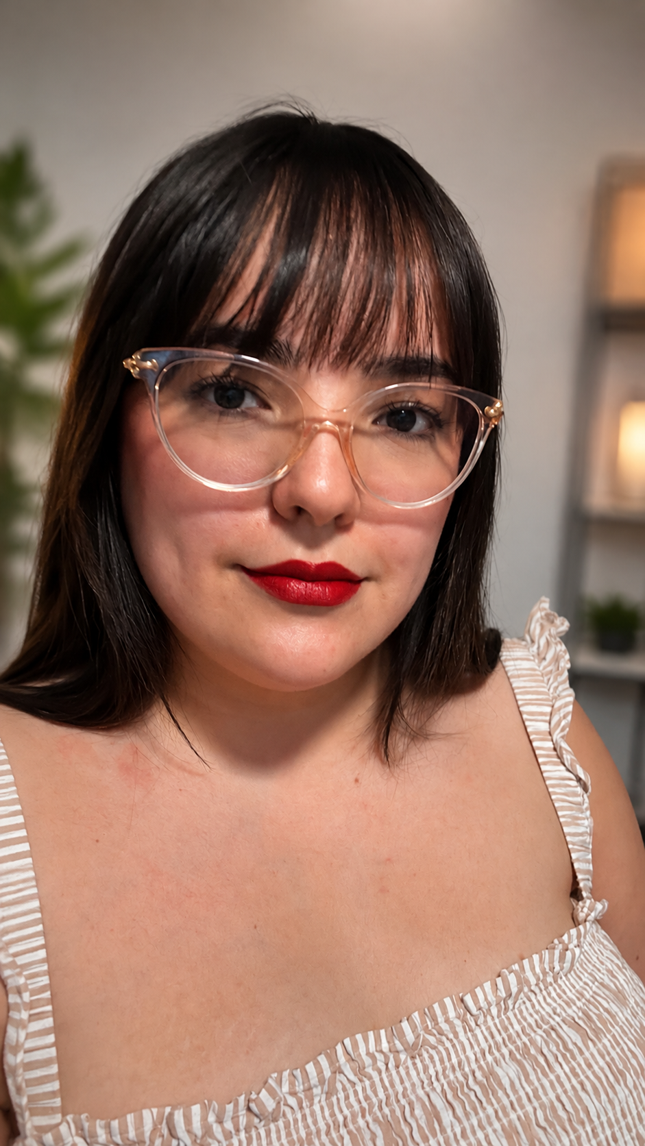 A woman with dark hair and bangs wearing clear cat-eye eyeglasses with gold metal frames and gold wire temples, paired with a cream and white striped top.