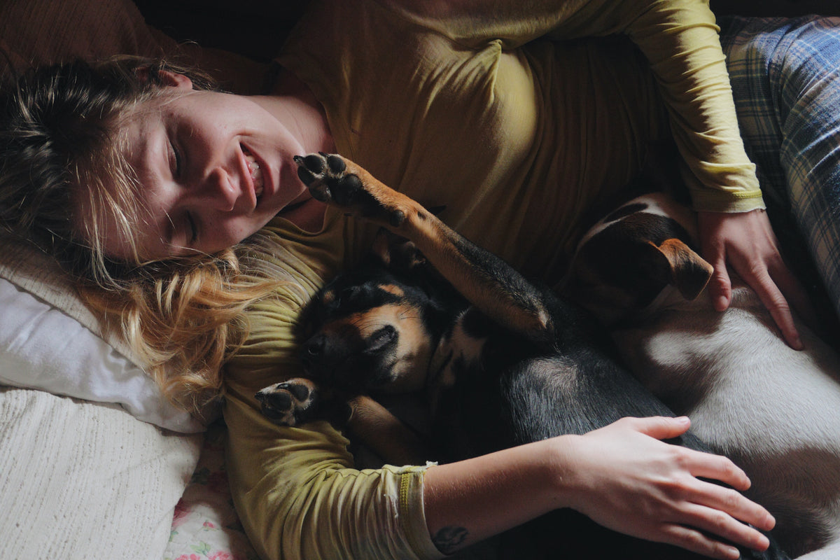 A woman lying on a bed with her eyes closed, smiling as she pets a black and tan dog and a white dog resting on her chest. She is wearing a yellow-green long-sleeve top and plaid pajama pants.